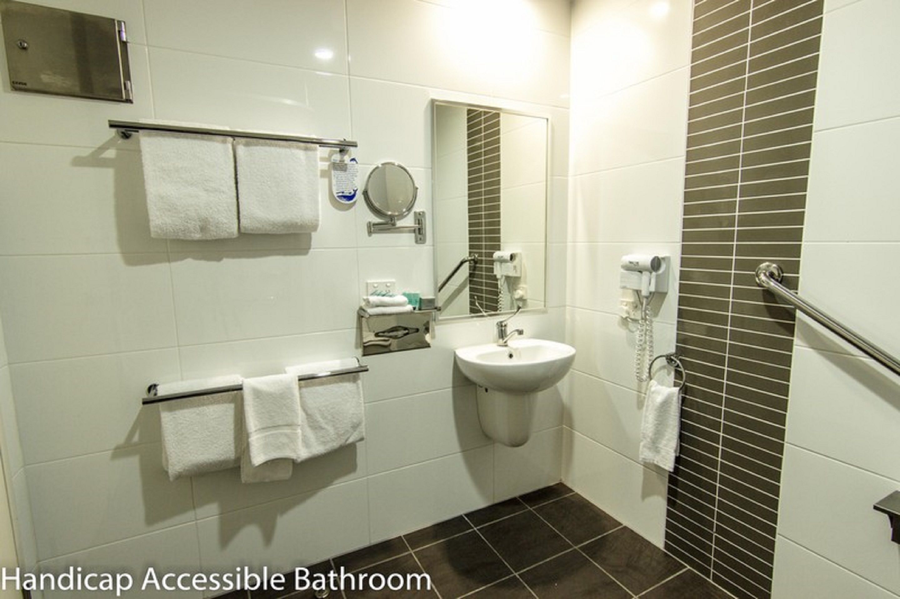 Une salle de bain adaptée aux personnes handicapées, avec des carreaux blancs, un lavabo, un miroir et un sèche-mains.