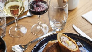 Une table à manger avec une bouteille de vin rouge dont on verse le contenu dans un verre, une assiette de nourriture et un bol de frites.