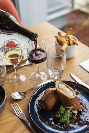 A dining table with a bottle of red wine being poured into a glass, a plate of food, and a bowl of fries. - ibis Lyon Sud Oullins (Oullins)