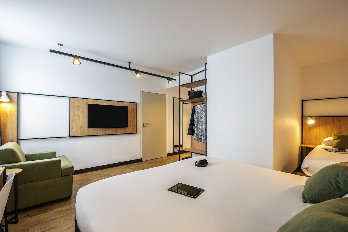 A modern hotel room with a large bed, a seating area with a sofa and chairs, a TV mounted on the wall, and a wardrobe with a mirror.