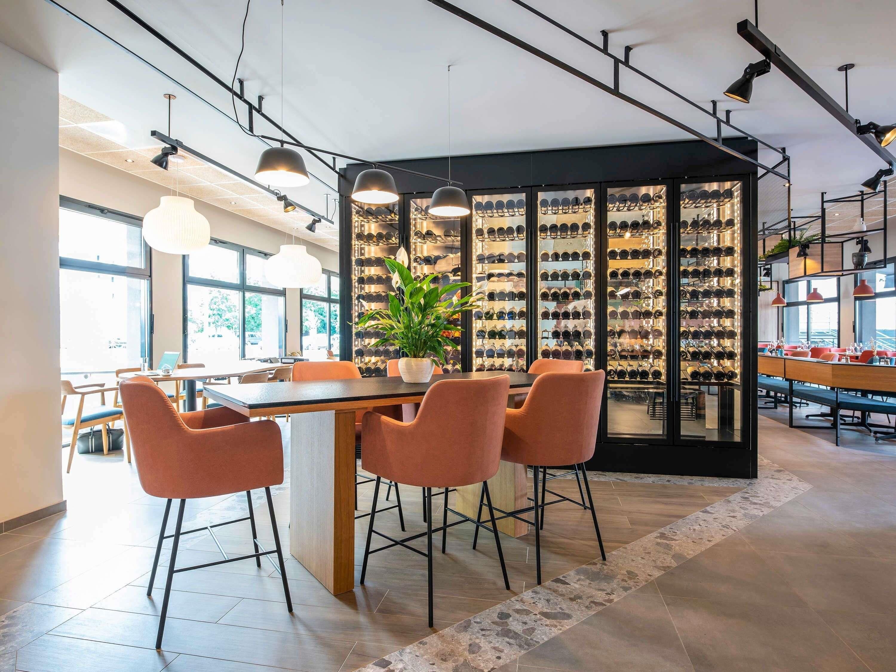 A modern interior with a dining area featuring a wooden table and orange chairs, a wine rack with bottles, and pendant lighting.