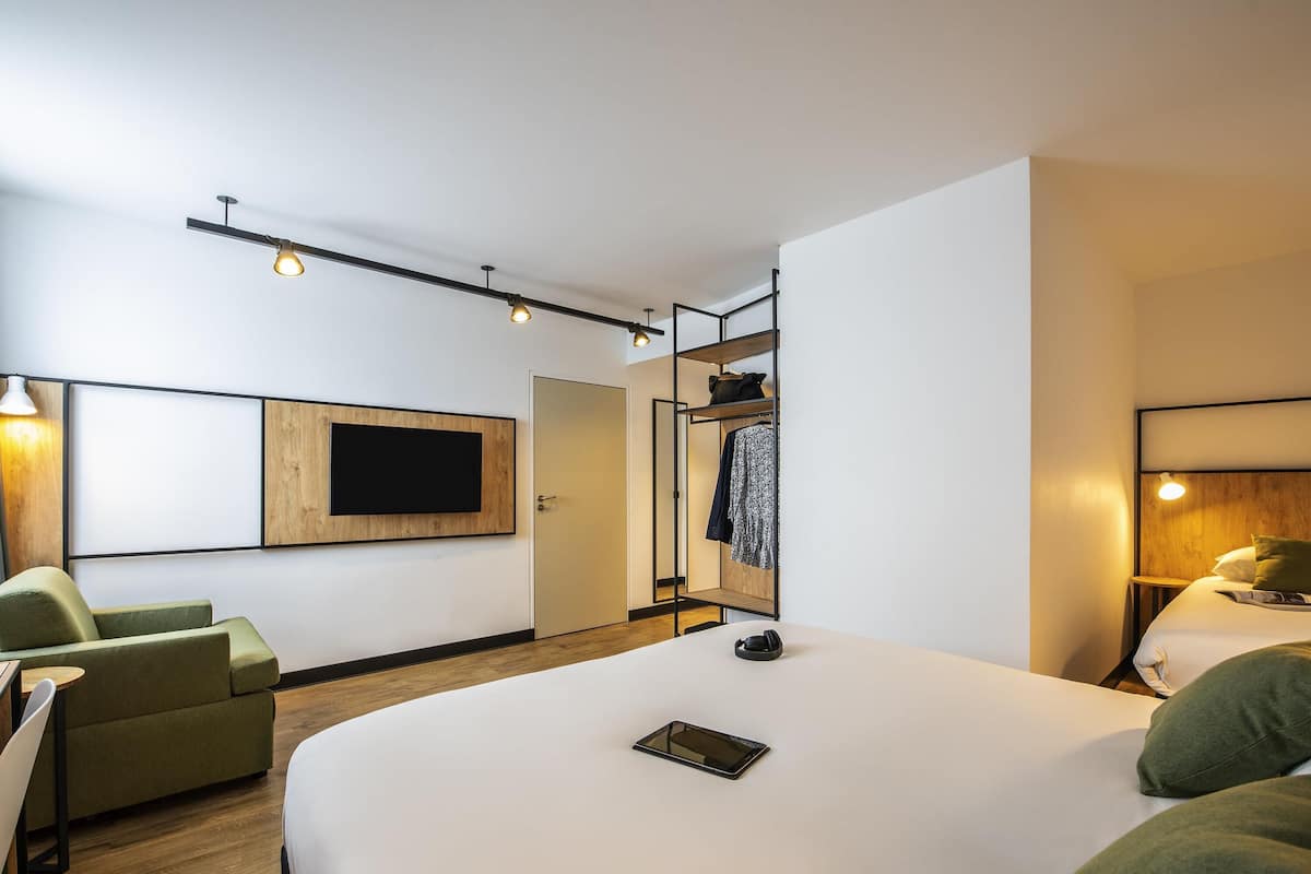 Quarto de hotel moderno com uma cama grande, área de estar com sofá e poltronas, televisão na parede e armário com espelho.