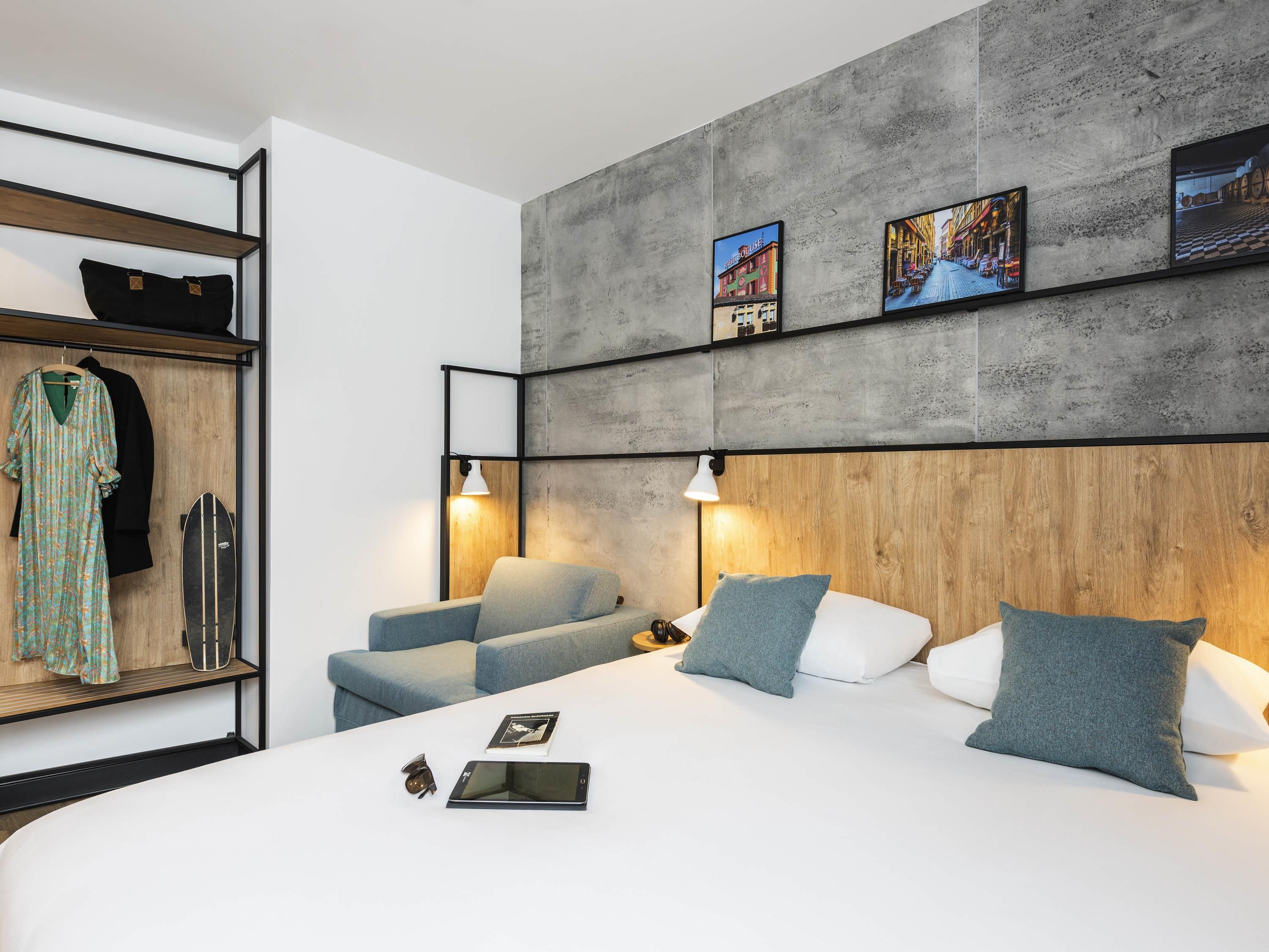 A modern hotel room with a large bed, a seating area, and a wardrobe with clothes and a surfboard.