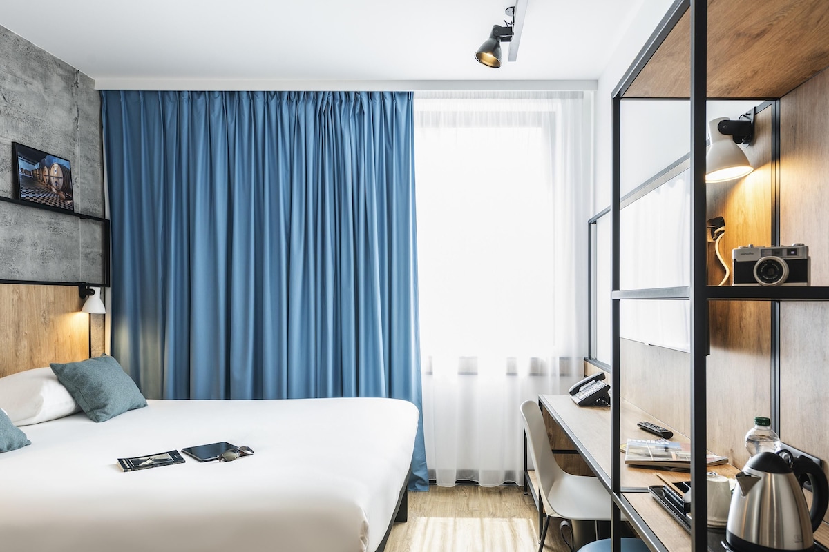 A modern hotel room with a bed, a desk, a chair, a kettle, a camera, and a window with curtains.