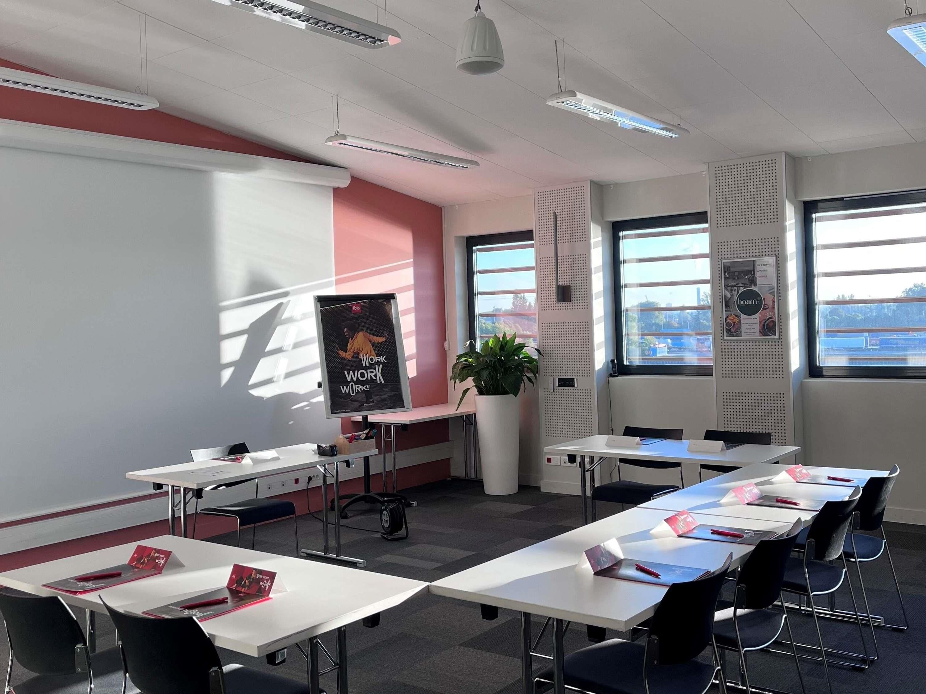 A modern conference room with white tables, black chairs, a large poster on the wall, and large windows allowing natural light.