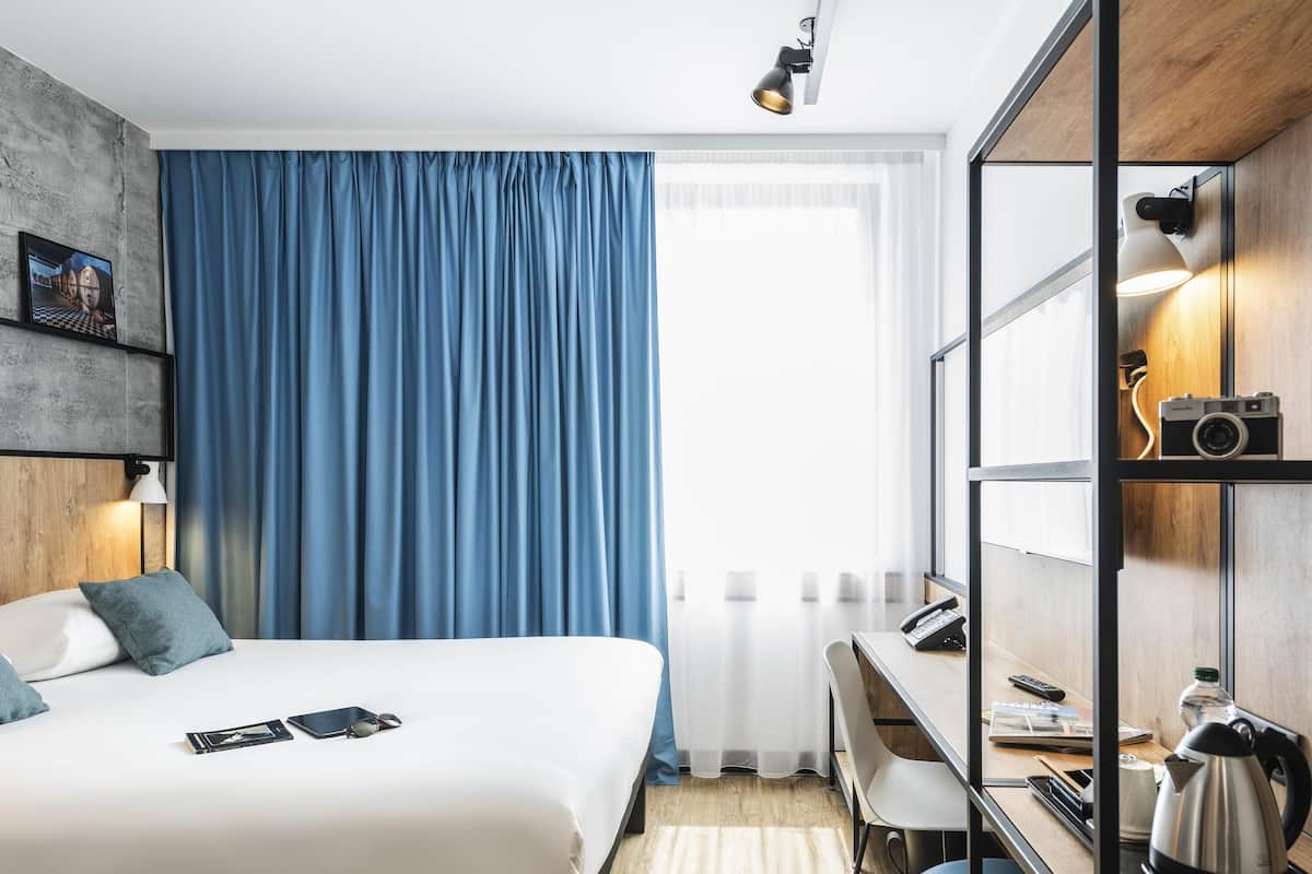 Quarto de hotel moderno com cama, escrivaninha, cadeira, chaleira, câmera e janela com cortinas.