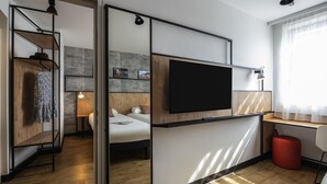 A modern hotel room with a large flat-screen TV, wooden flooring, a sliding door closet, and a red ottoman. - ibis Lyon Sud Oullins (Oullins)