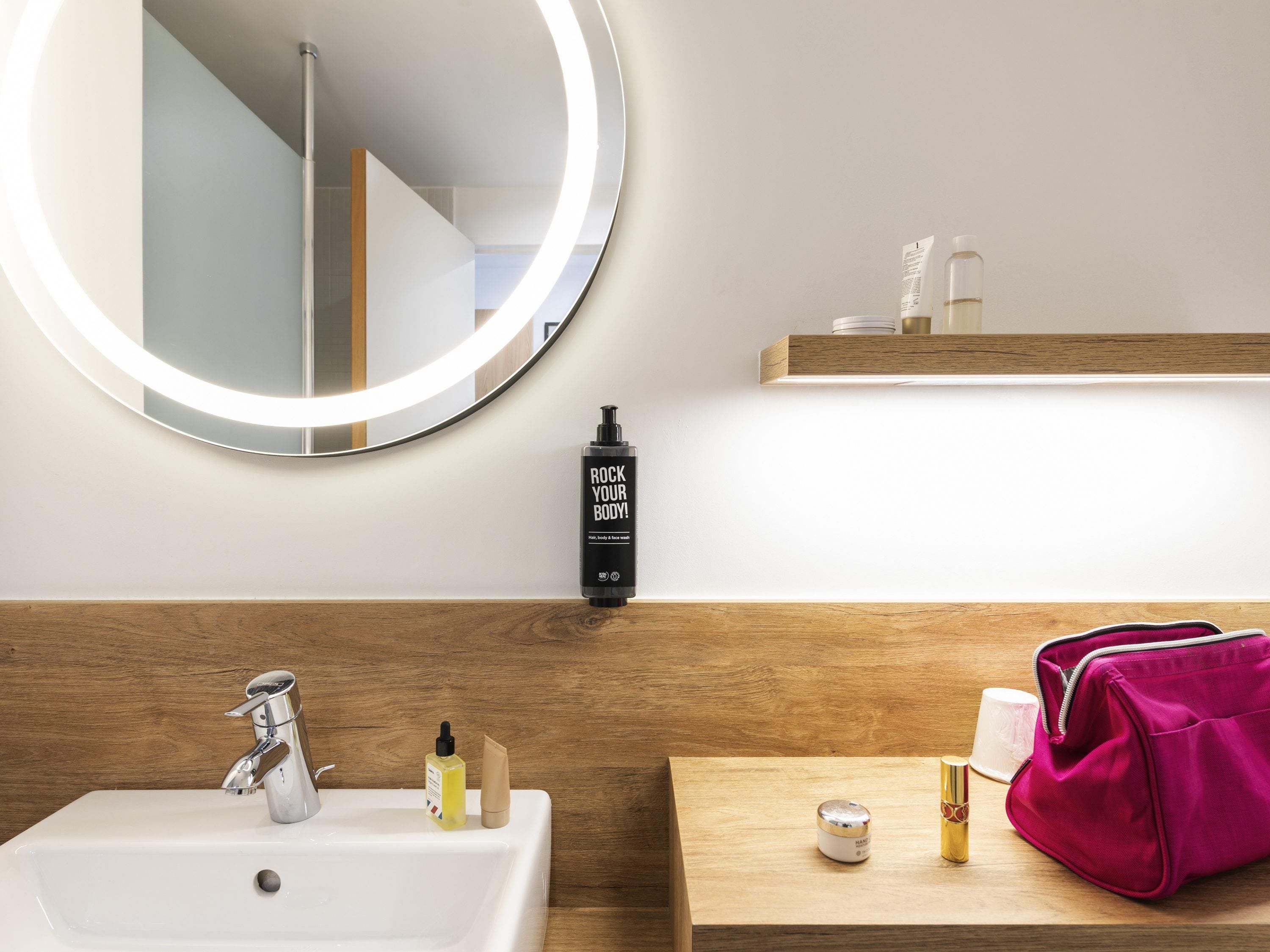 Ein Badezimmer mit weißem Waschbecken, einem Spiegel mit runder Beleuchtung und einer Holzkommode mit Pflegeprodukten.