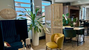 A modern interior with a blue sofa, a yellow chair, a large potted plant, a wooden partition, and a dining area with green chairs. - ibis Lyon Sud Oullins (Oullins)