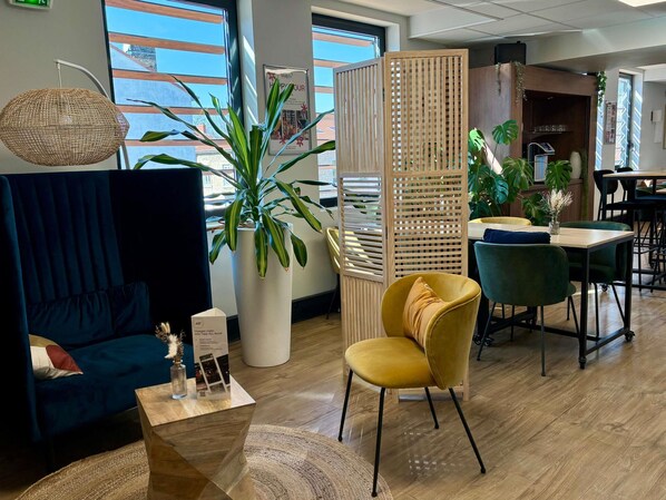 Ein modernes Interieur mit einem blauen Sofa, einem gelben Sessel, einer großen Topfpflanze, einer Holzvertäfelung und einem Essbereich mit grünen Stühlen.