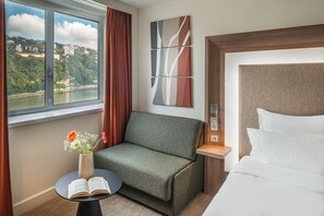 A hotel room with a bed, a sofa, a small table with an open book and flowers, a window with a view of a river and buildings, and a wall with a piece of abstract art.