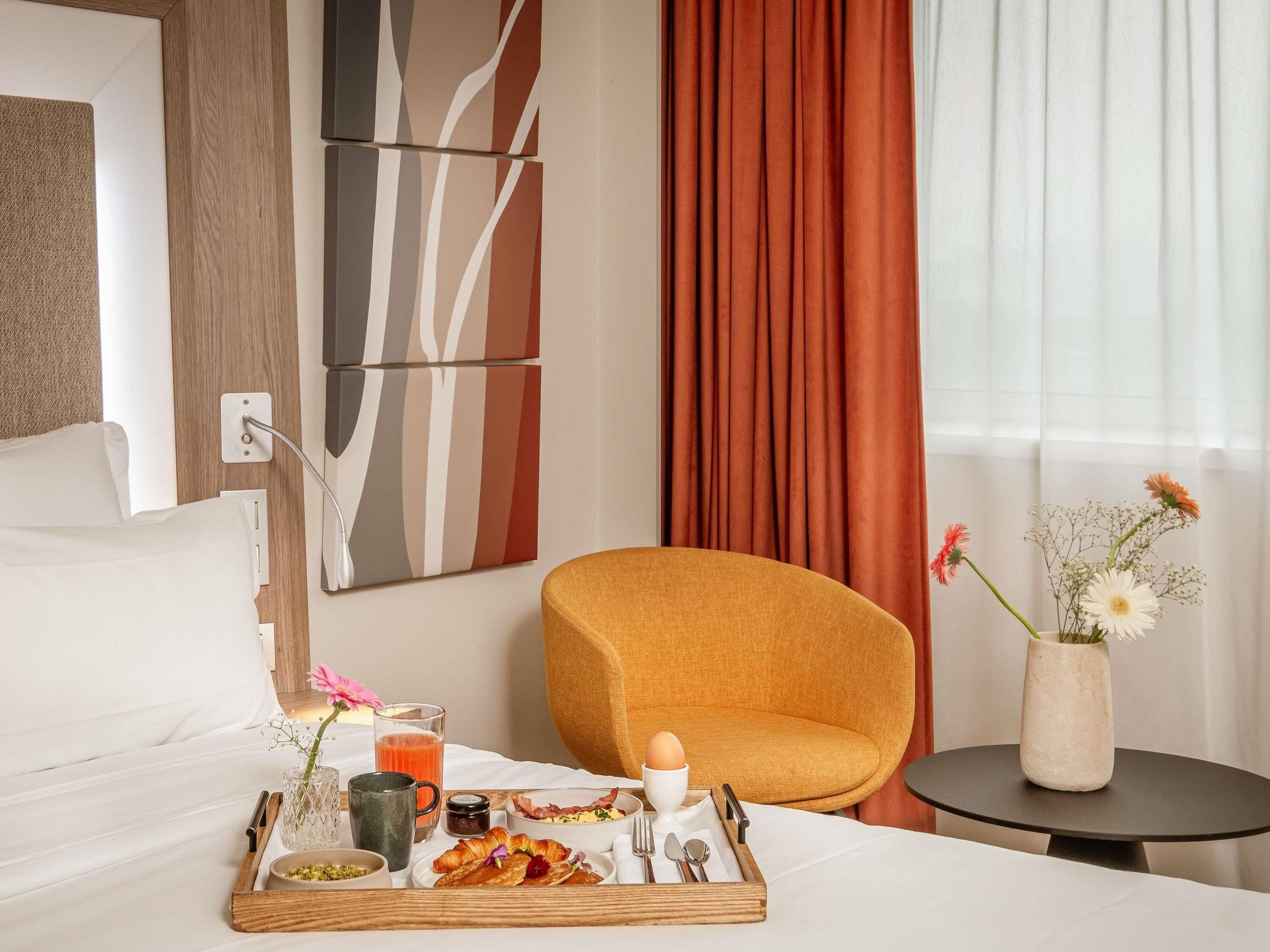 Hotelkamer met een bed, een ontbijttafel, een stoel, een vaas met bloemen en een muur met abstracte kunst.
