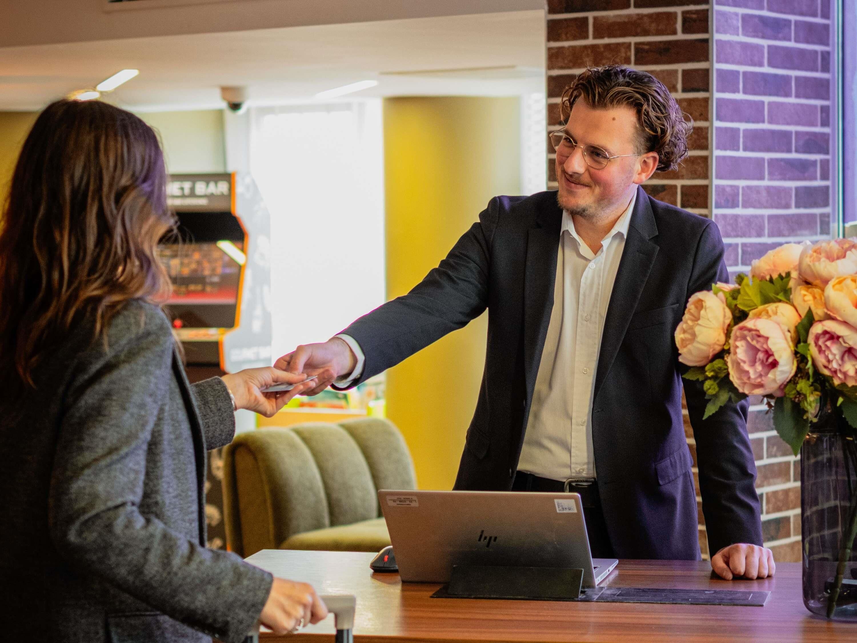 A man in a suit handing a credit card to a woman at a reception desk with a laptop and a bouquet of flowers.