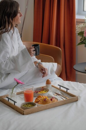 Una persona sentada en una cama con una bandeja de desayuno, que incluye croissants y un vaso de zumo de naranja.