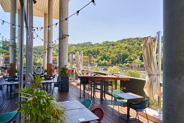 A terrace with tables and chairs, a view of a river, and a hilly landscape in the background. - Novotel Lyon Centre Confluence Bord de Saone (Lyon)