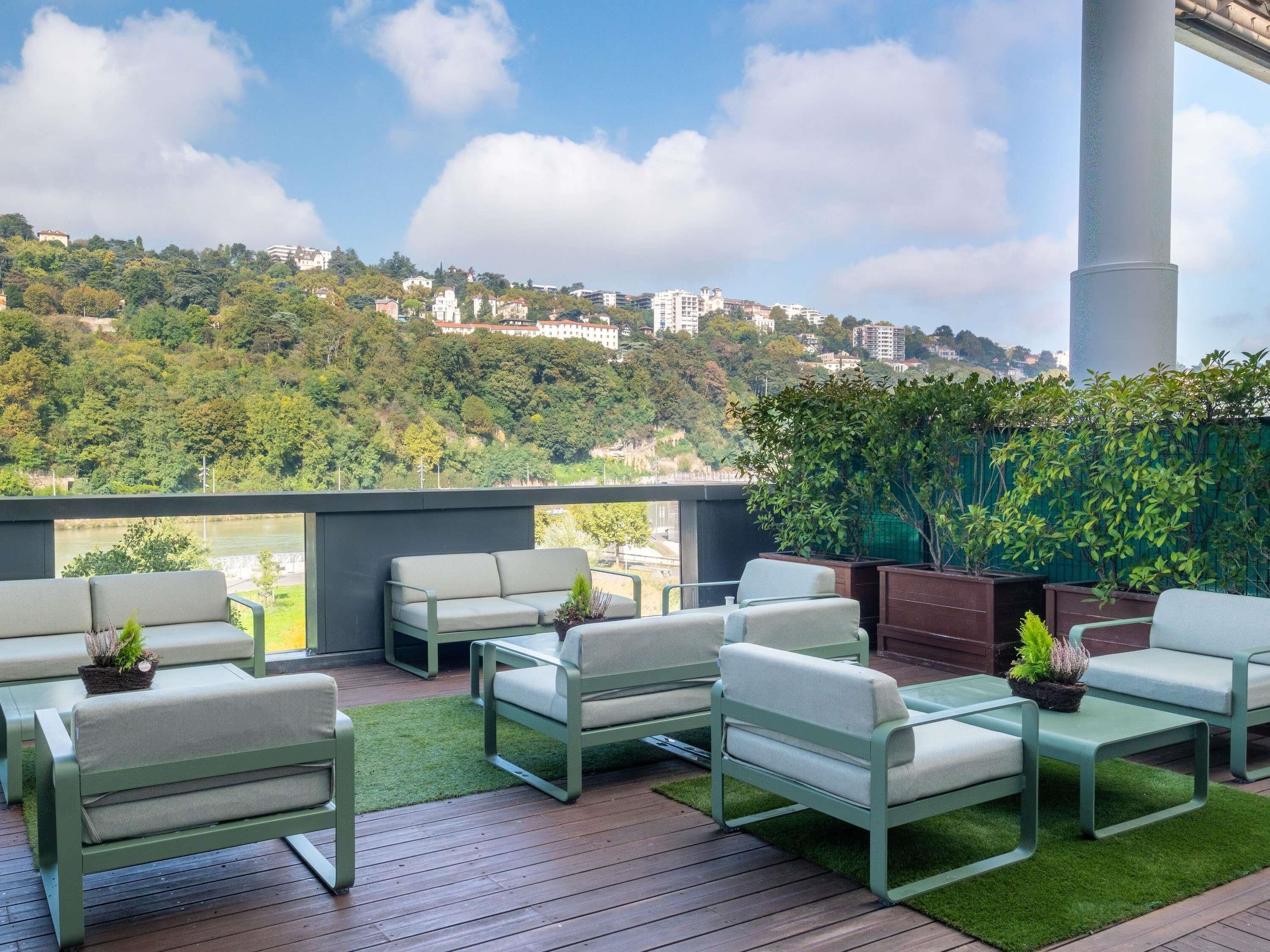 A rooftop terrace with outdoor seating, a view of a hillside, and a large cylindrical structure.