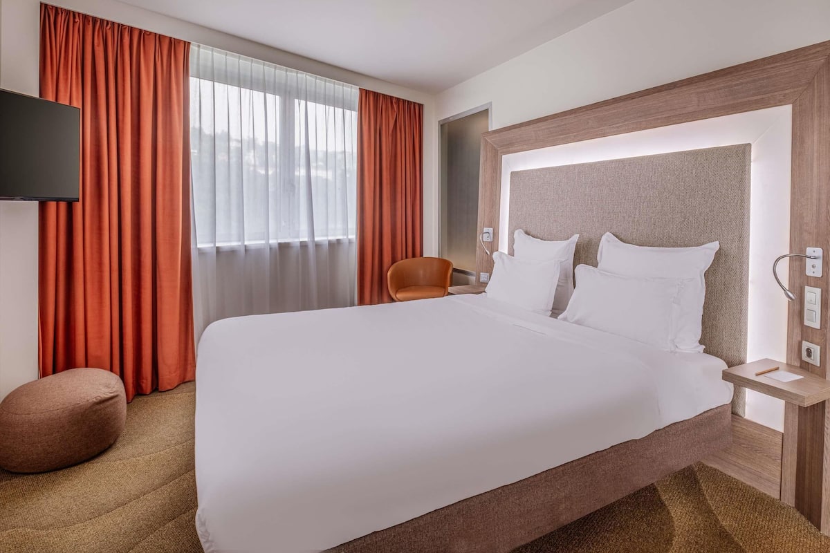 Habitación de hotel con una cama grande, televisión, una silla y ventana con cortinas.