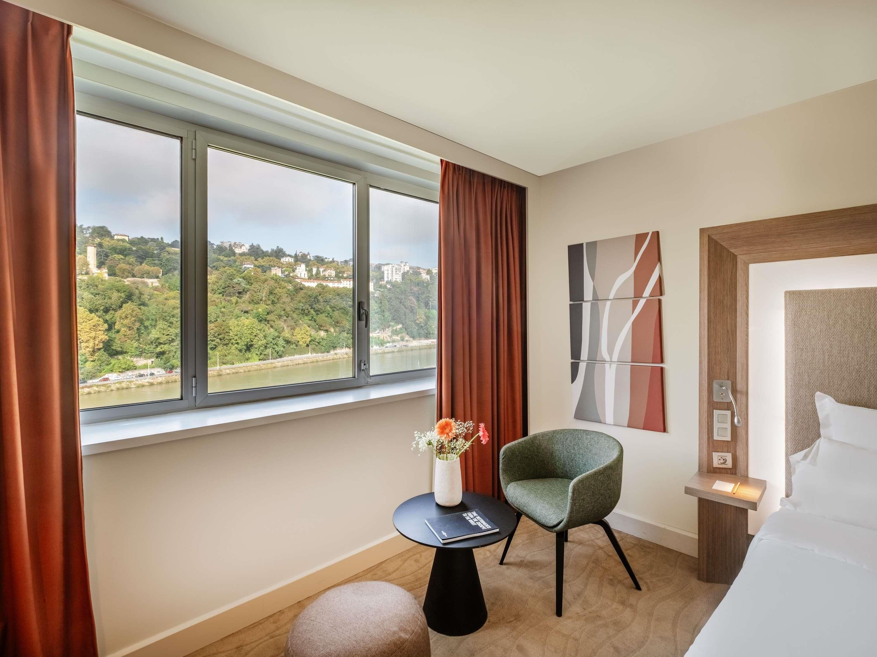 Habitación de hotel con un ventanal grande, una silla verde, una mesita con un jarrón de flores y una cama con ropa blanca.