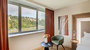 Habitación de hotel con un ventanal grande, una silla verde, una mesita con un jarrón de flores y una cama con ropa blanca.