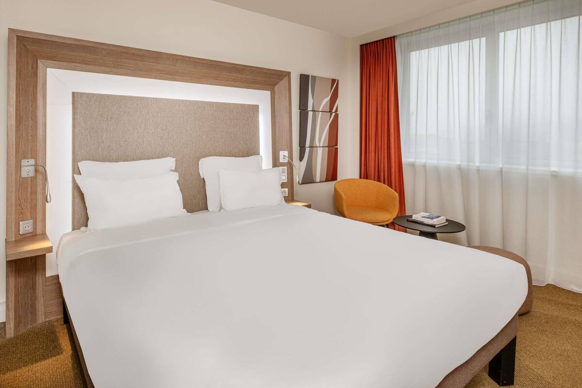 Habitación de hotel con una cama grande, una mesita redonda pequeña, una silla y una ventana con cortinas.