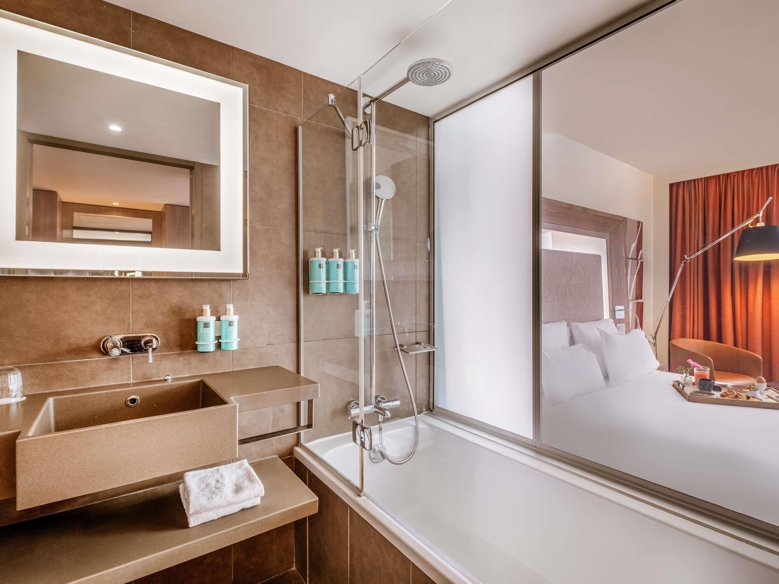 A modern hotel bathroom with a large mirror, a shower, a sink with a towel, and toiletries.