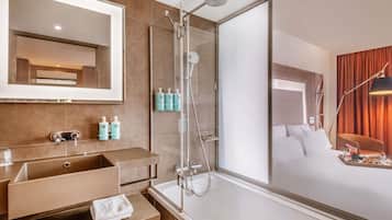 A modern hotel bathroom with a large mirror, a shower, a sink with a towel, and toiletries.