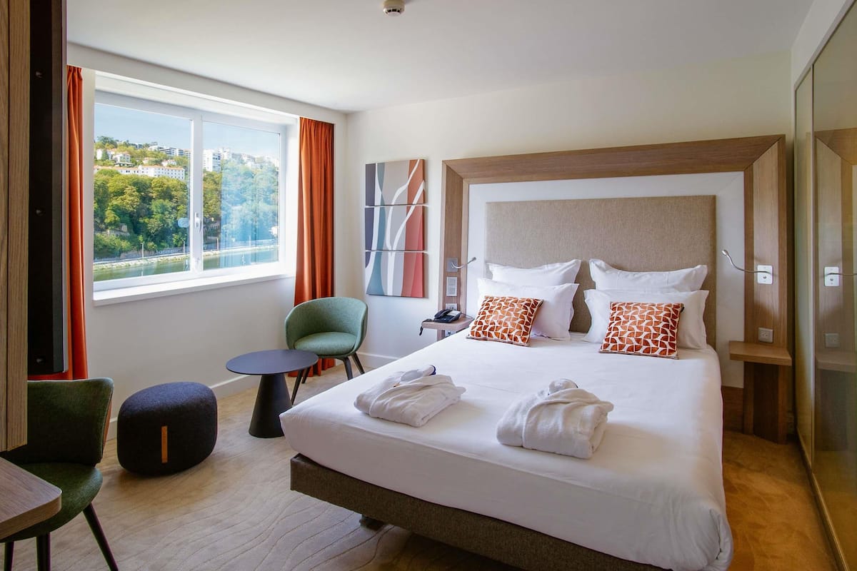 Habitación de hotel con una cama, una silla, una mesita y vistas a un río y edificios.