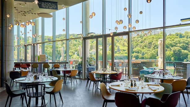 Un restaurante con varias mesas redondas y sillas, ventanas amplias que dan a vistas de zonas verdes y edificios, y lámparas esféricas colgantes.