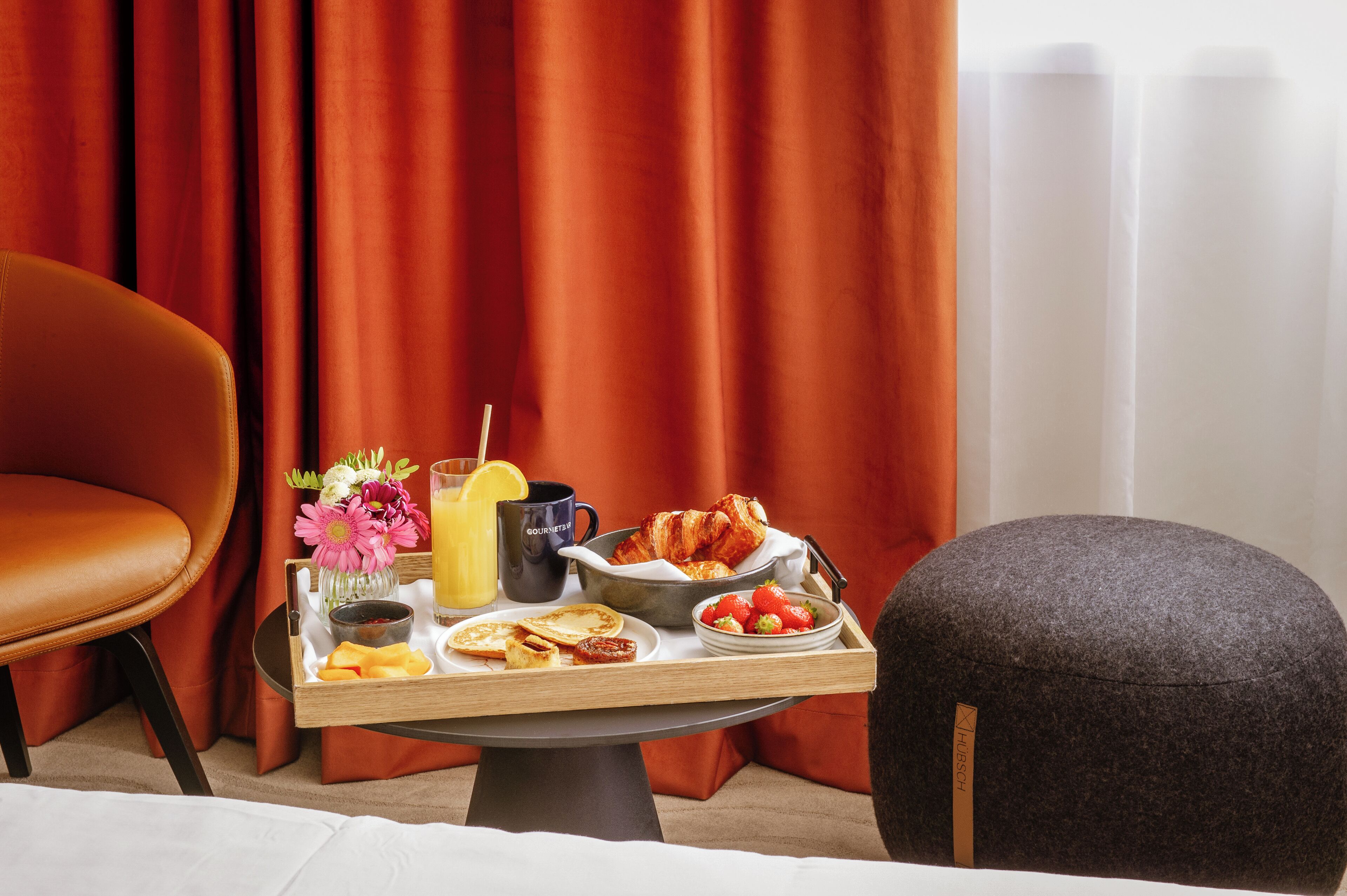 Ein Frühstückswagen mit Croissants, Erdbeeren, Orangensaft und einer Blumenvase auf einem Tisch in einem Zimmer mit roten Vorhängen und einem Ledersessel.