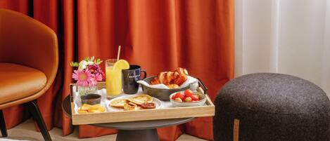 Ein Frühstückswagen mit Croissants, Erdbeeren, Orangensaft und einer Blumenvase auf einem Tisch in einem Zimmer mit roten Vorhängen und einem Ledersessel.