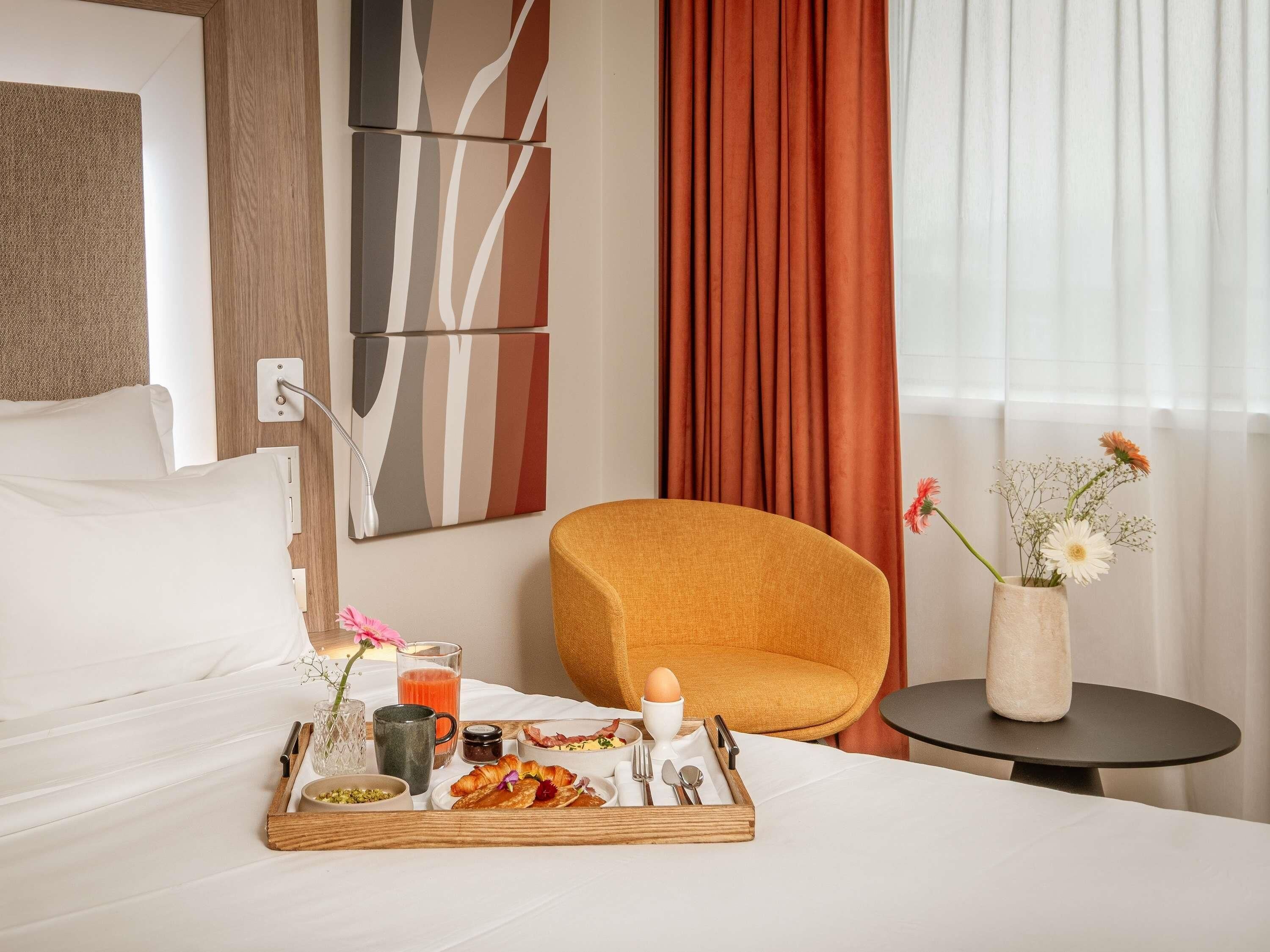 Ein Hotelzimmer mit Bett, Frühstückswagen, Stuhl, Blumen in einer Vase und einem Fenster mit Vorhängen.