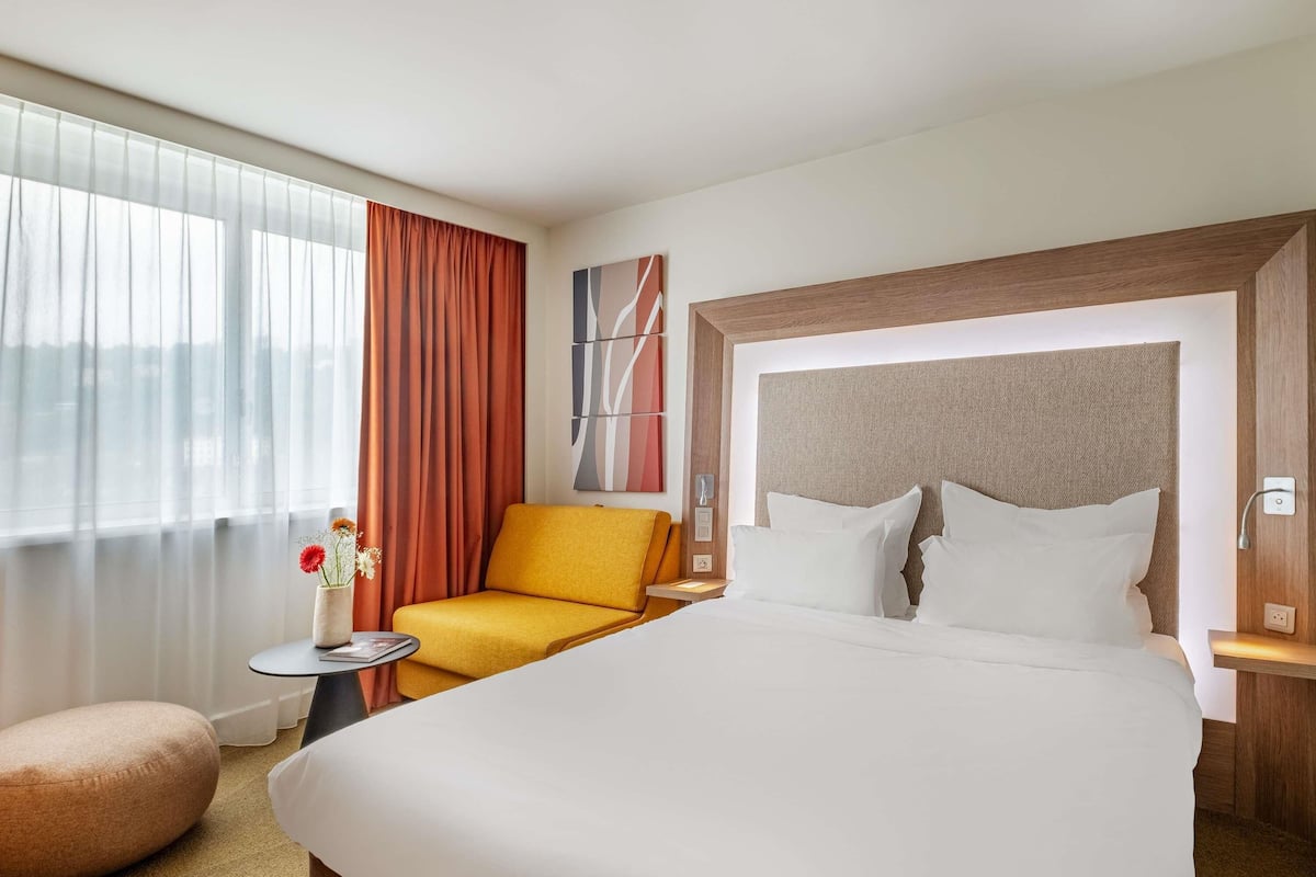 Un cuarto de hotel con una cama grande, un sillón amarillo, una mesita redonda con un florero y vistas de la ciudad a través de la ventana.