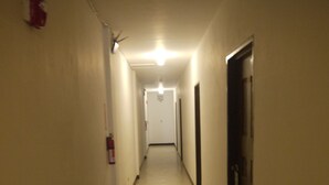 A narrow, well-lit hallway with a fire extinguisher on the left, a door on the right, and a ceiling light at the end. - Himawari Hotel (Saipan)