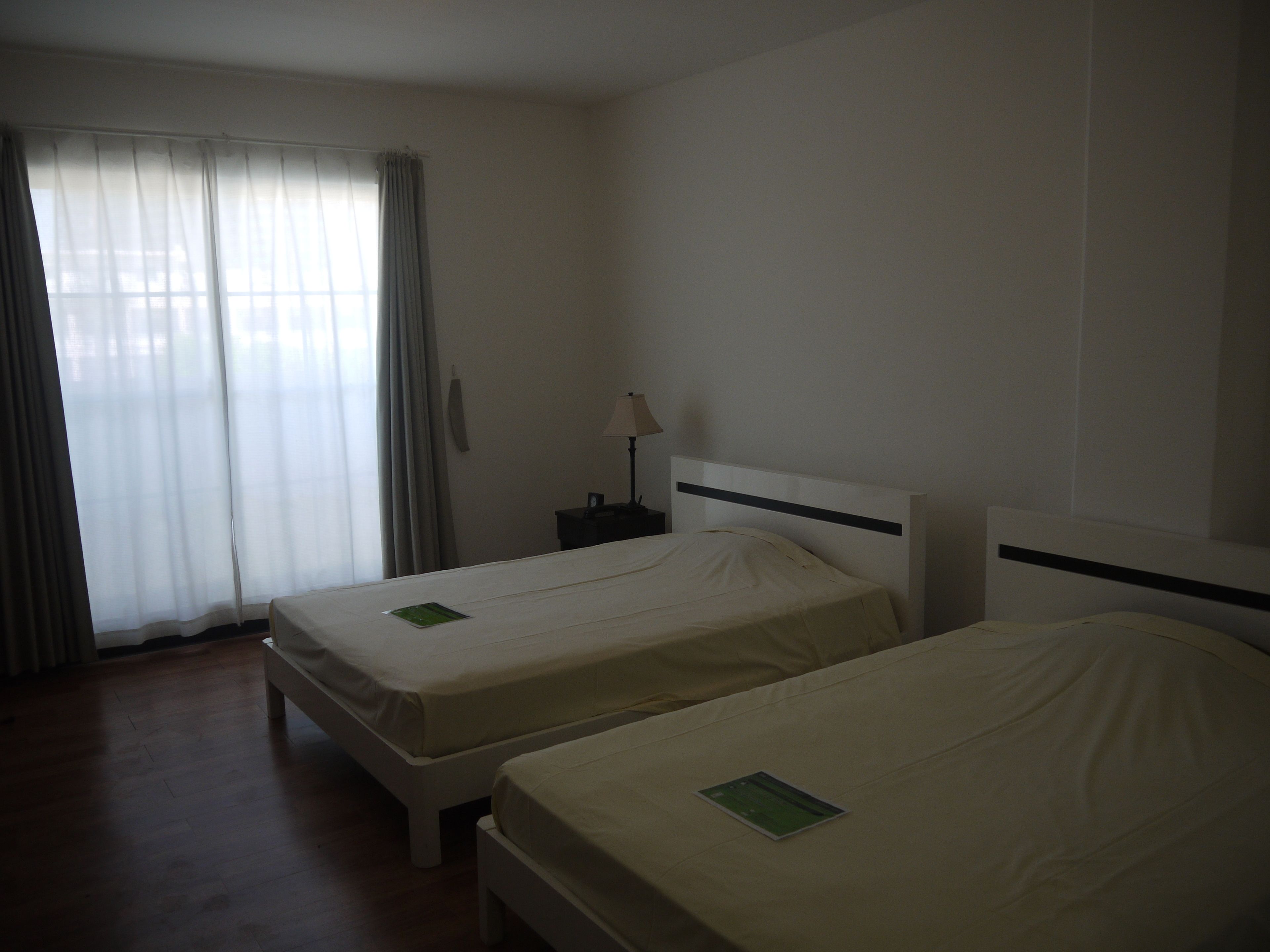 Een hotelkamer met twee enkele bedden, een nachtkastje met een lamp en een raam met dunne gordijnen.
