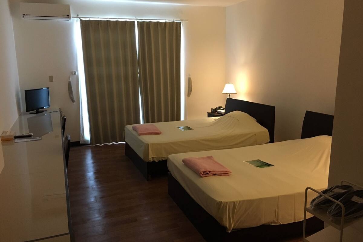 Habitación de hotel con dos camas, un escritorio con computadora, una lámpara y una ventana con cortinas.