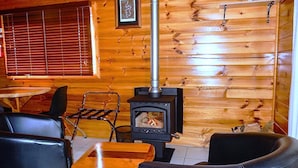 A cozy cabin interior with a wood-burning stove, wooden walls, a dining table, and leather chairs. - Lumera Chalets (St Marys)