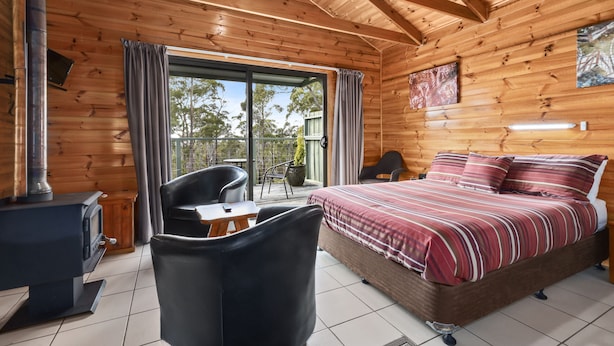 A wooden cabin room with a bed, a table, a chair, and a wood stove. - Lumera Chalets (St Marys)
