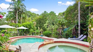 Una piscina con dos sillas de descanso y sombrillas, rodeada de abundante follaje y palmeras.