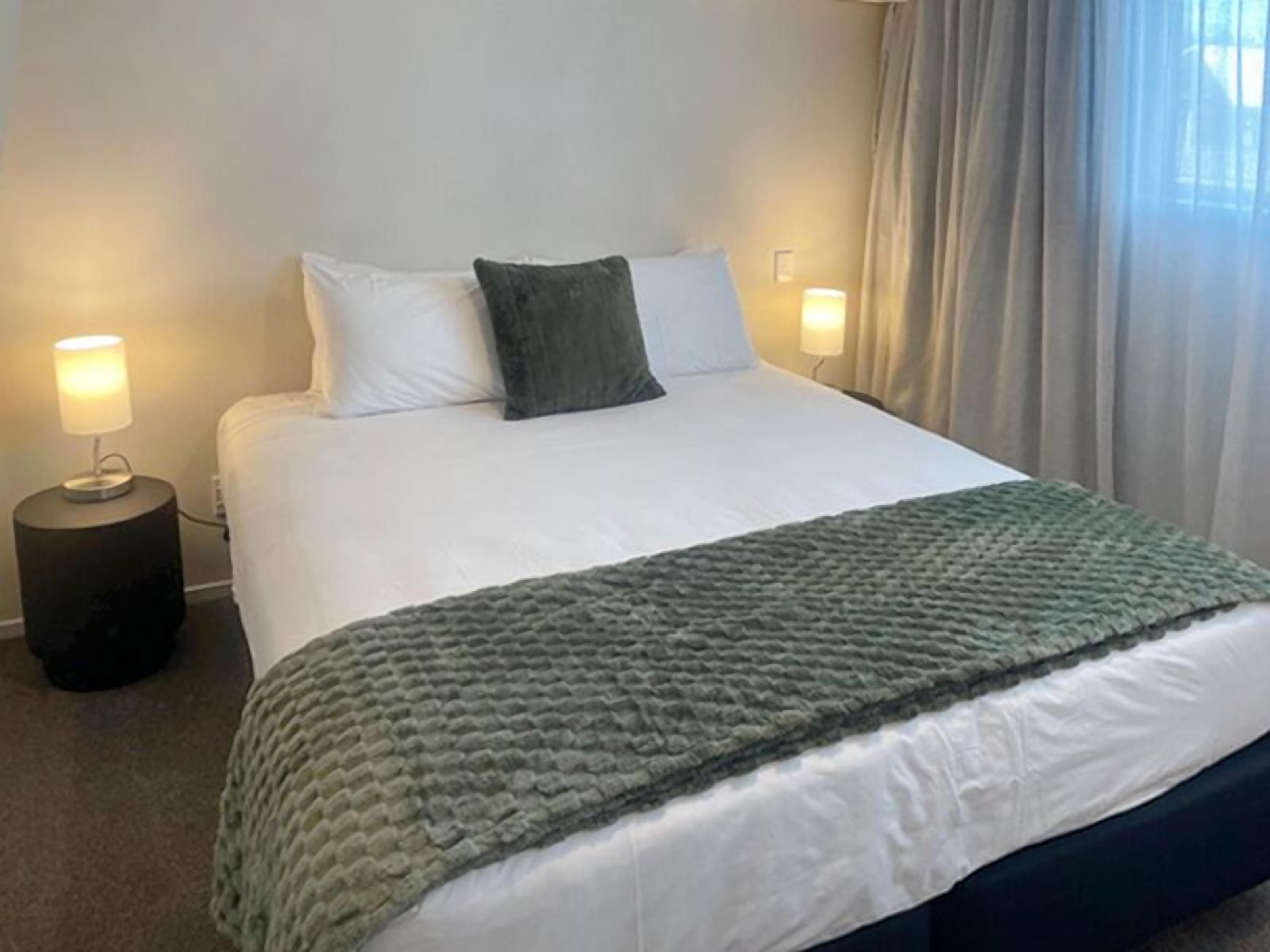 A neatly made bed with white linens and a green knitted throw, two bedside lamps, and a window with curtains.