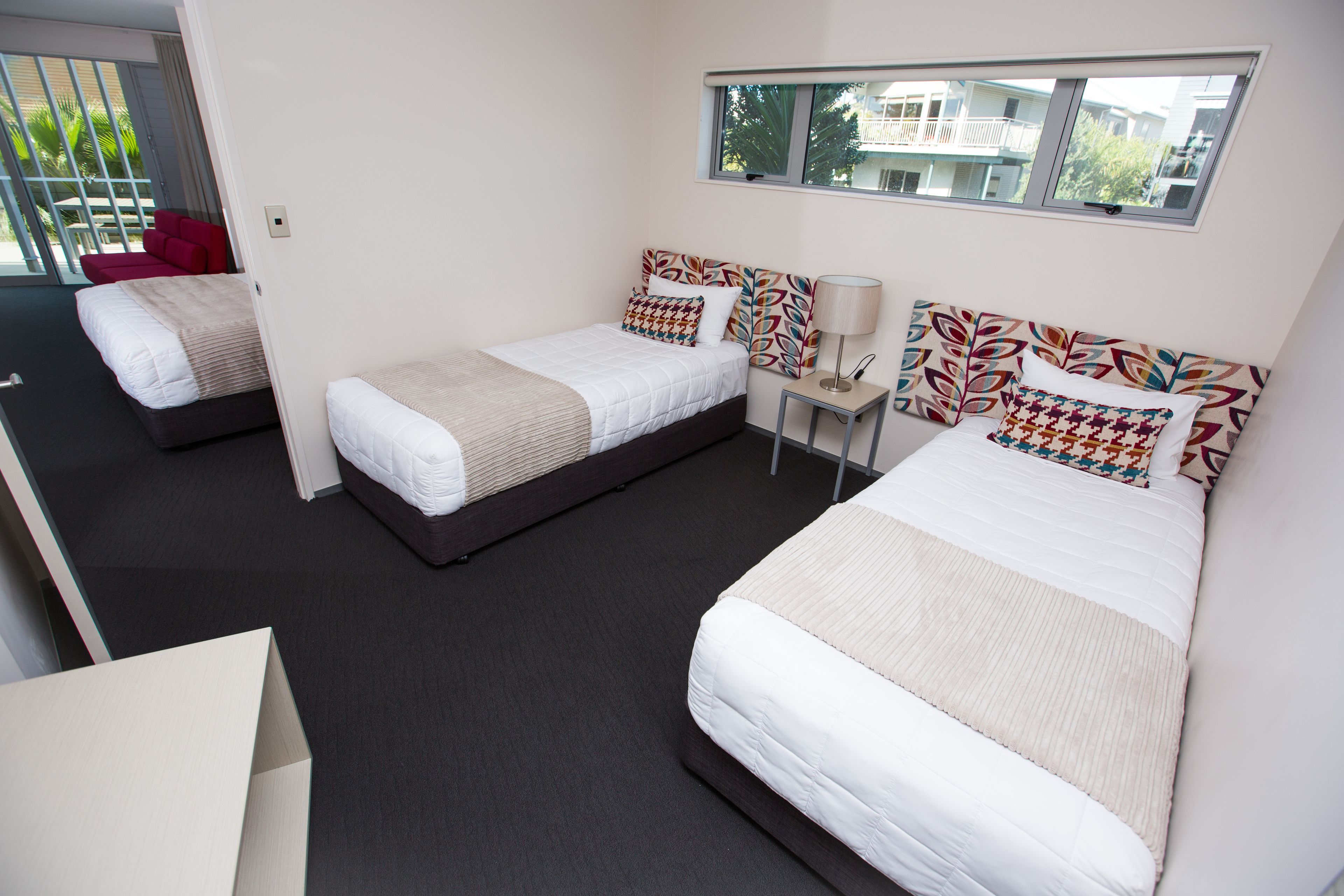 Habitación de hotel con tres camas, una mesita de noche con lámpara y ventana con vista a edificios.