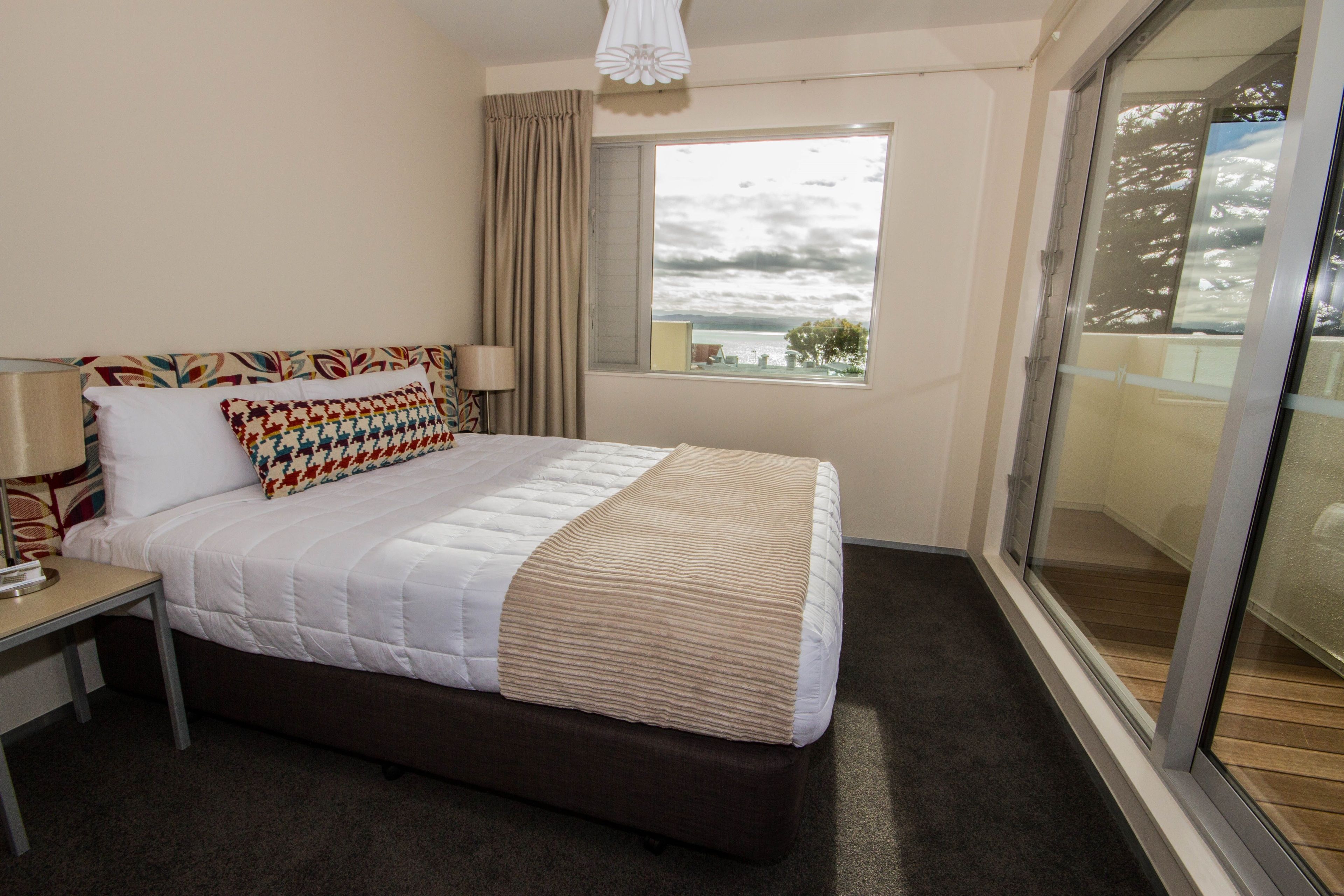 A hotel room with a bed, a nightstand, a window with a view, and a sliding glass door.