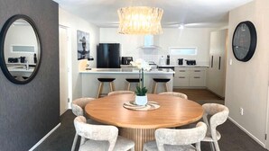 A modern kitchen with a central island, a dining area with a round table and six chairs, and a large mirror on the left wall.