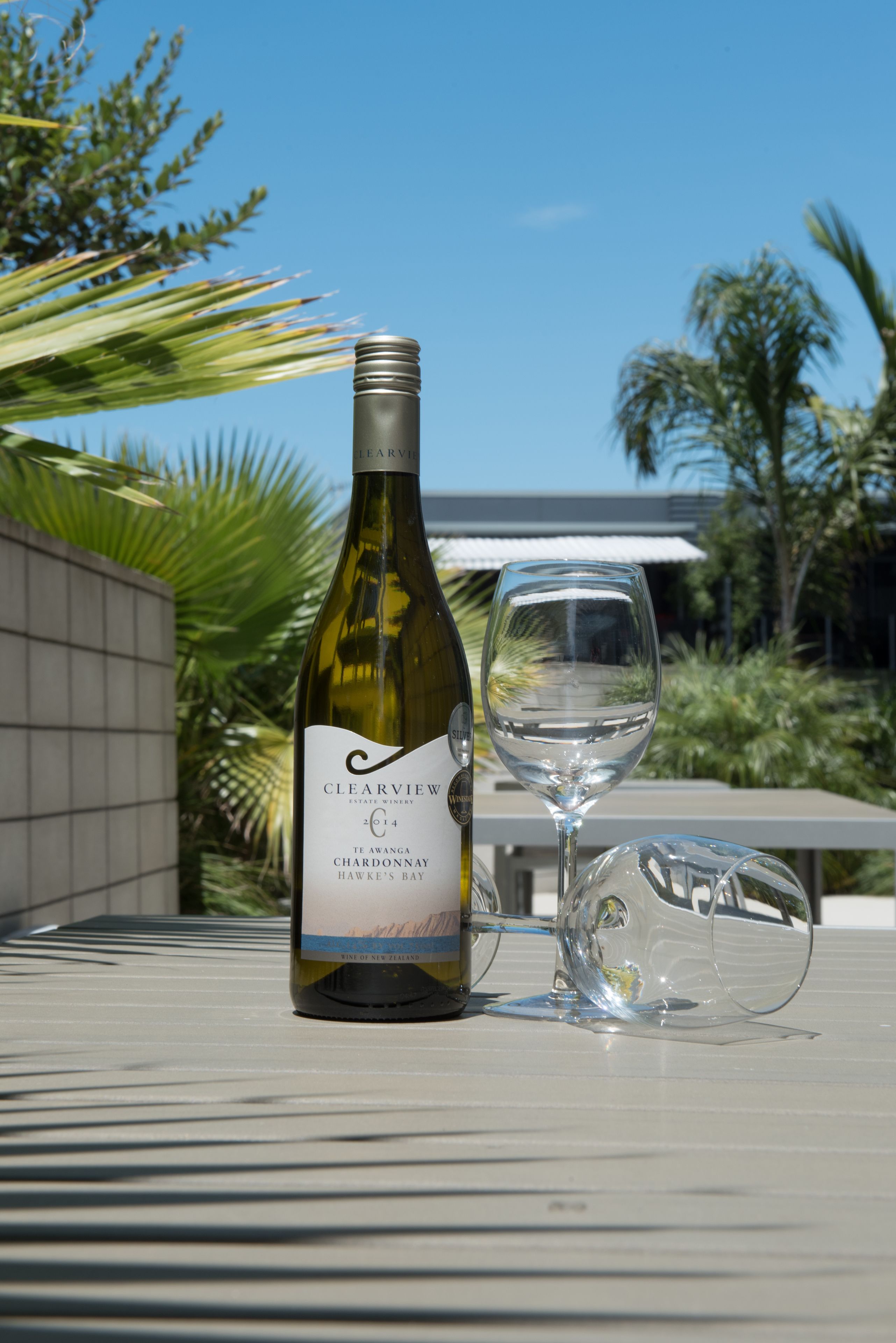 Uma garrafa de vinho e um copo sobre uma mesa ao ar livre, com um edifício e vegetação ao fundo.