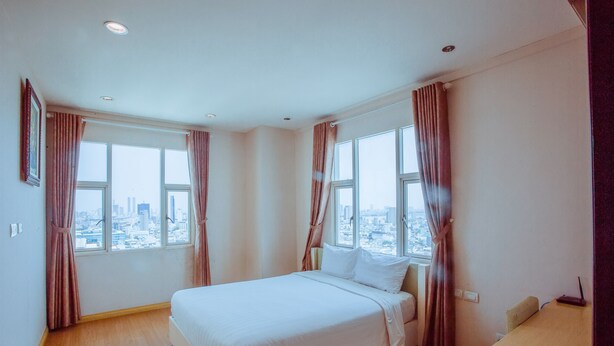 A hotel room with a large bed, two windows with curtains, a desk, and a view of the city.