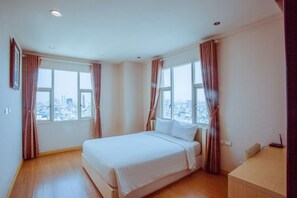Habitación de hotel con una cama grande, dos ventanas con cortinas, un escritorio y vistas a la ciudad.