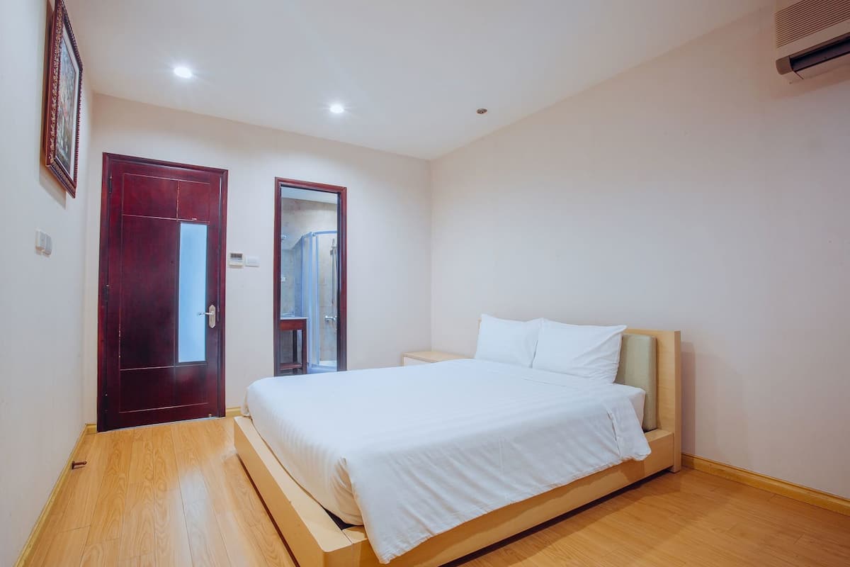 A bedroom with a wooden floor, a bed with white bedding, a wooden door, and a mirror.