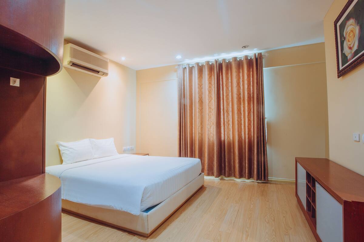 A hotel room with a bed, wooden flooring, an air conditioning unit, a curtain, and a framed picture on the wall.