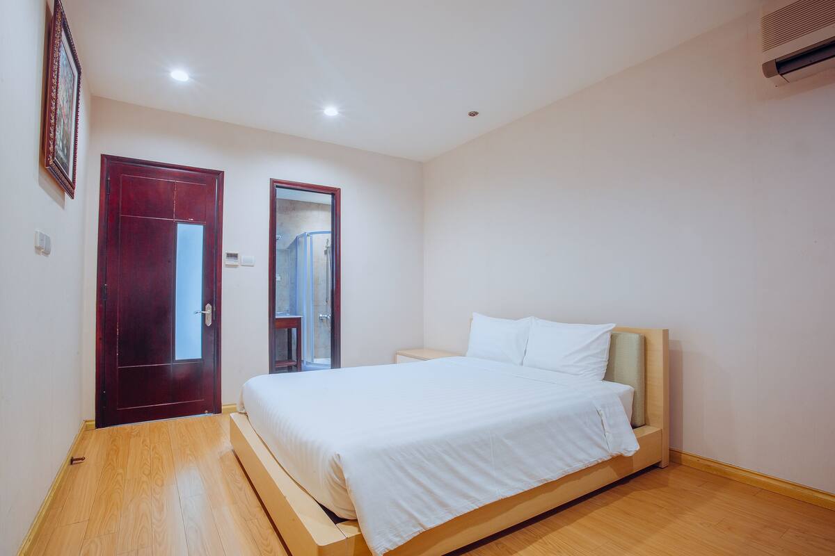 A bedroom with a wooden floor, a bed with white bedding, a nightstand, a door, and a wall-mounted air conditioner.