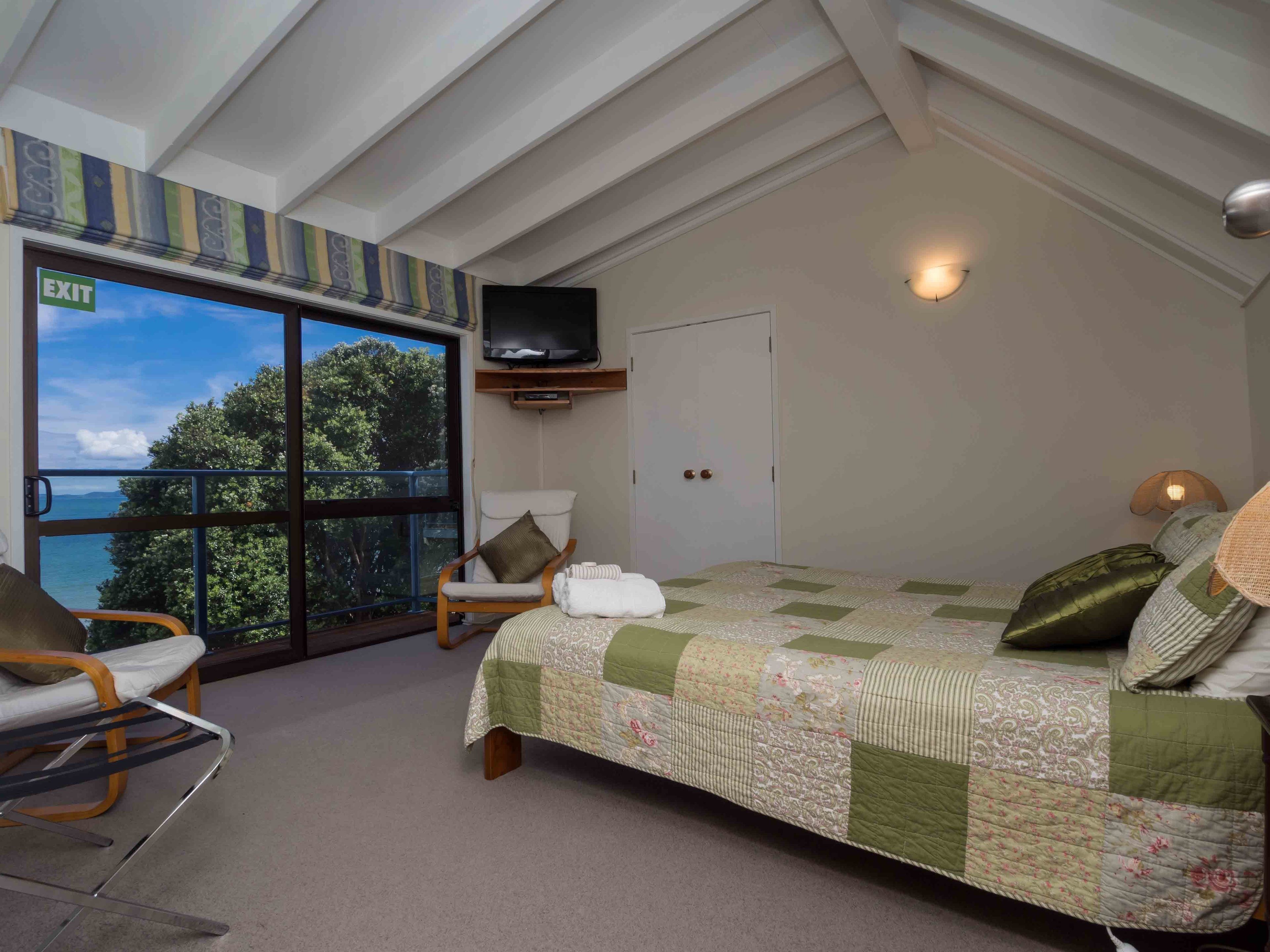 A bedroom with a large window offering a view of trees and a body of water, a bed with a quilt, a chair, and a television mounted on the wall.