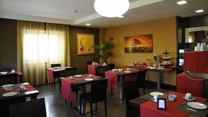 Espaço de refeições de hotel com mesas preparadas para o pequeno-almoço, um quadro grande na parede e uma luminária pendente moderna.